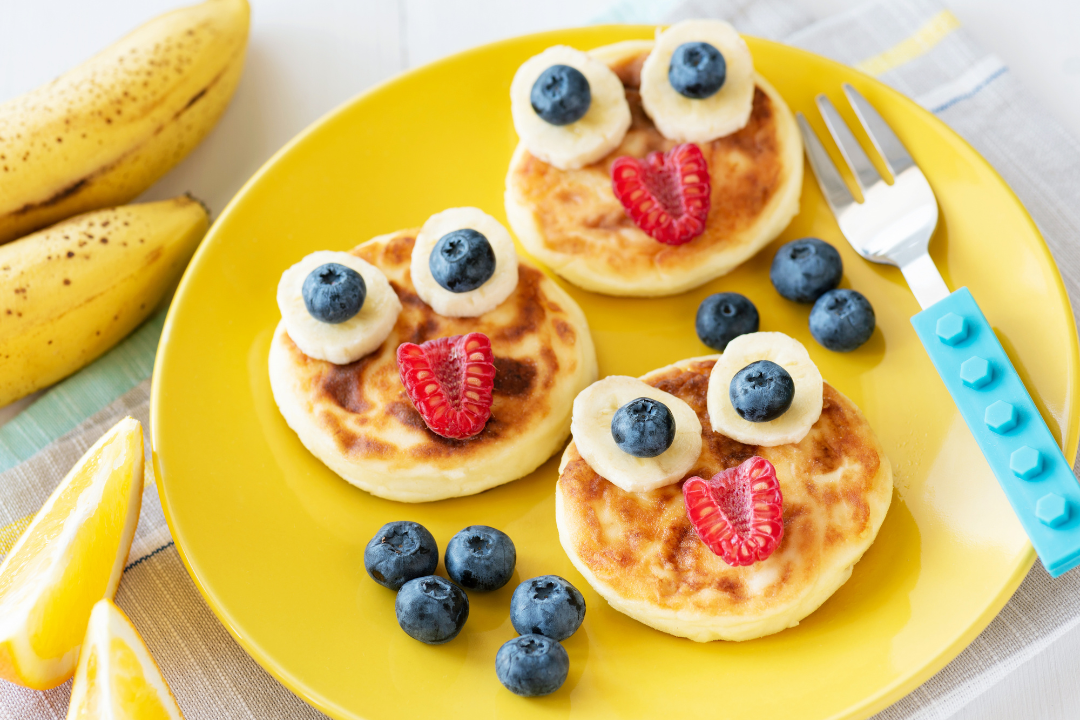 Three fiber-rich smiley face fruit pancakes and a child's fork on a plate with bananas and lemon slices on the side